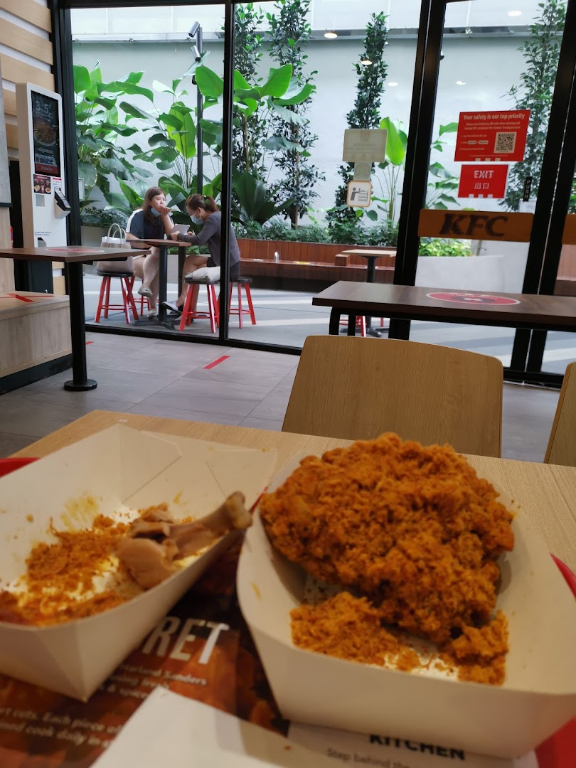 A close-up of KFC fried chicken on a table, with diners and a mall view in the background.