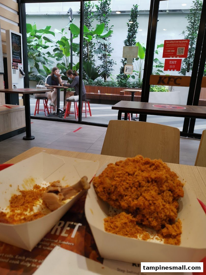 A close-up of KFC fried chicken on a table, with diners and a mall view in the background.