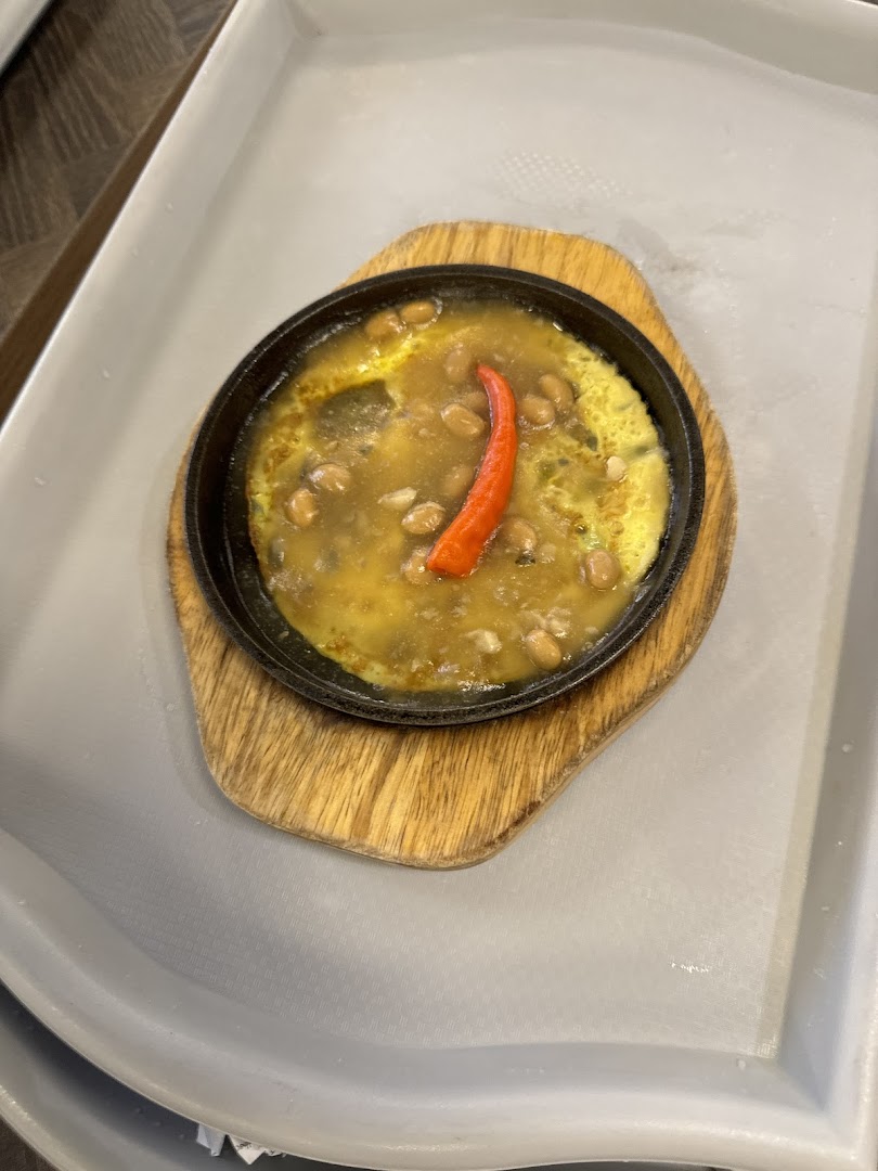 A sizzling hot plate dish with beans, a rich sauce, and a vibrant red chili, served on a wooden board.