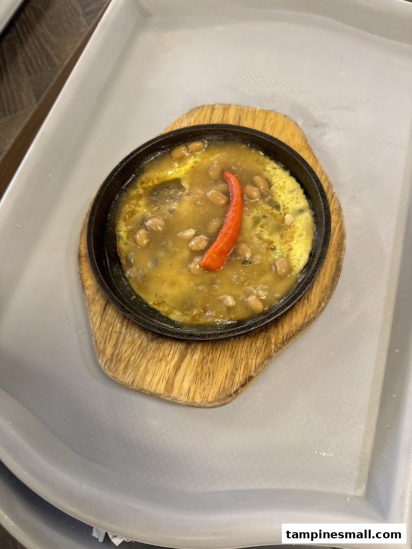 A sizzling hot plate dish with beans, a rich sauce, and a vibrant red chili, served on a wooden board.