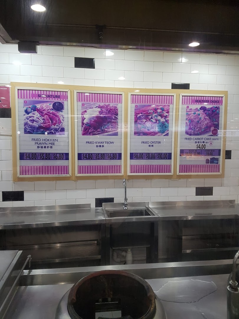 Menu display at Kopitiam featuring fried noodle and cake dishes with prices above a kitchen counter.
