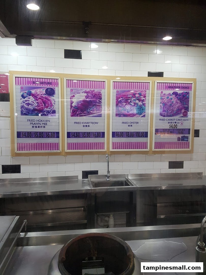 Menu display at Kopitiam featuring fried noodle and cake dishes with prices above a kitchen counter.