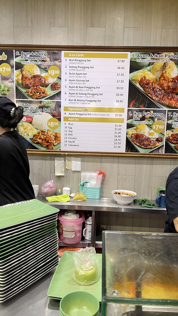 A food stall's menu board displays various set meals and add-ons, with a staff member in the foreground.