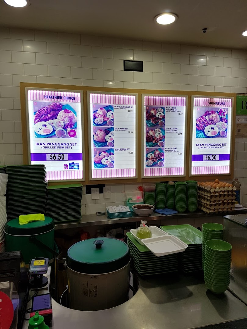 A food stall counter at Kopitiam featuring illuminated menu boards and stacked green dishes.