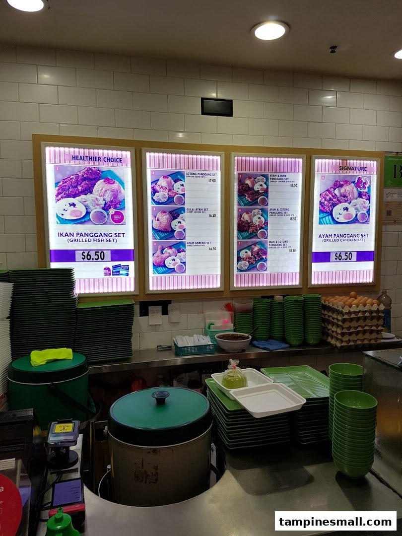 A food stall counter at Kopitiam featuring illuminated menu boards and stacked green dishes.