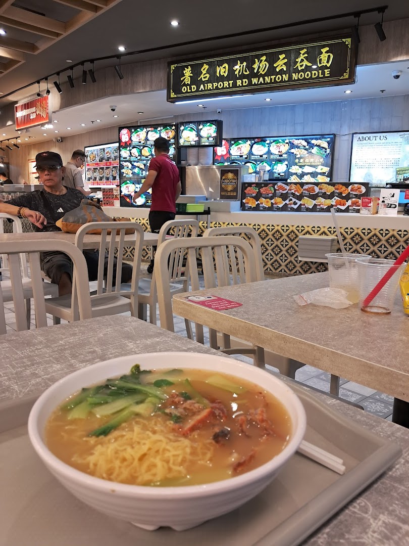 A delicious bowl of wanton noodles on a table in a lively Kopitiam food court setting.