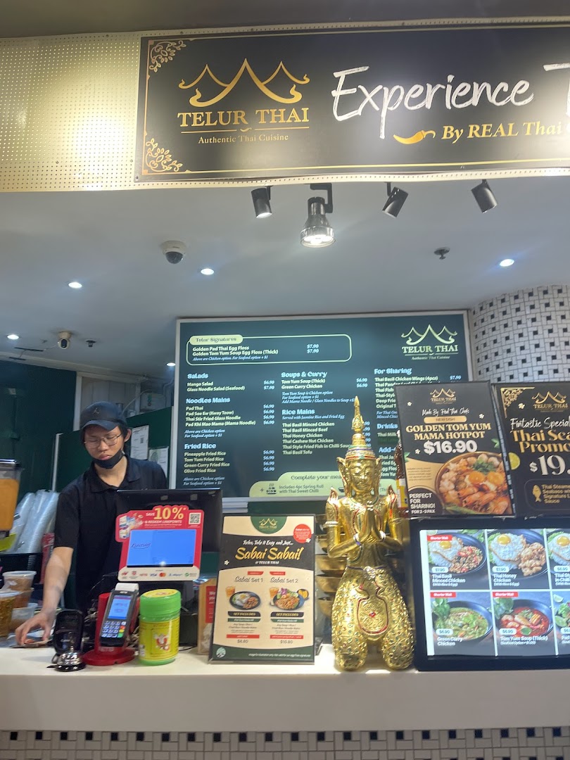 A staff member stands behind the counter of Telur Thai, featuring a large menu board and promotional displays.