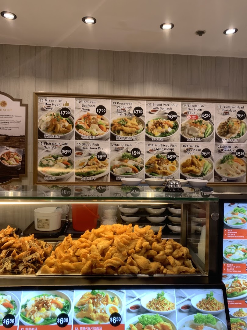 A food stall counter at Kopitiam featuring a large menu board displaying various fish and pork soup dishes, alongside trays of fried food.