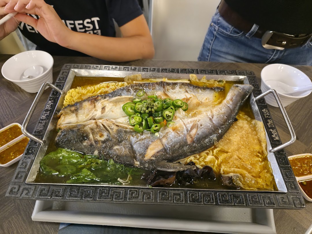 A large, flavorful whole fish dish served on a hot plate with noodles, vegetables, and sauces.
