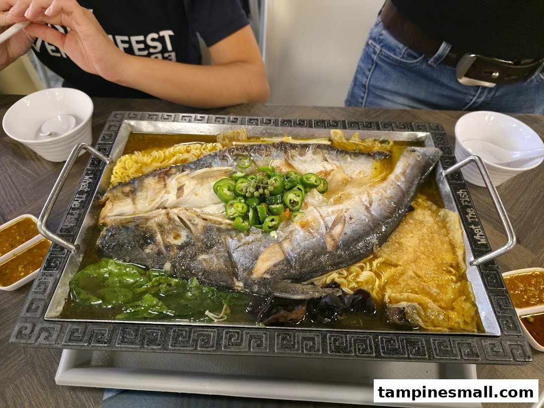 A large, flavorful whole fish dish served on a hot plate with noodles, vegetables, and sauces.