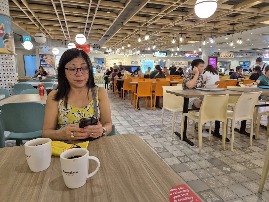A woman sits at a table in a bustling Kopitiam food court, with other diners and stalls in the background.