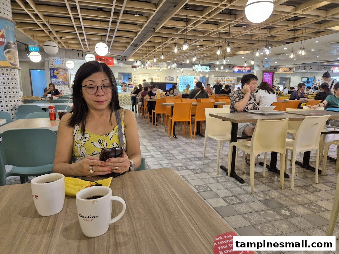 A woman sits at a table in a bustling Kopitiam food court, with other diners and stalls in the background.