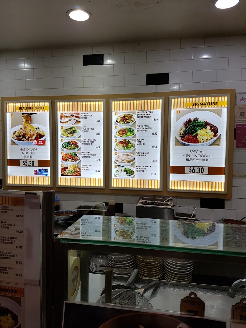 A brightly lit menu board at Kopitiam showcasing a variety of noodle dishes and prices above a food counter.