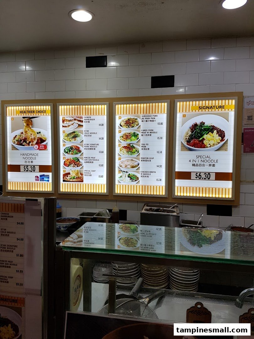 A brightly lit menu board at Kopitiam showcasing a variety of noodle dishes and prices above a food counter.