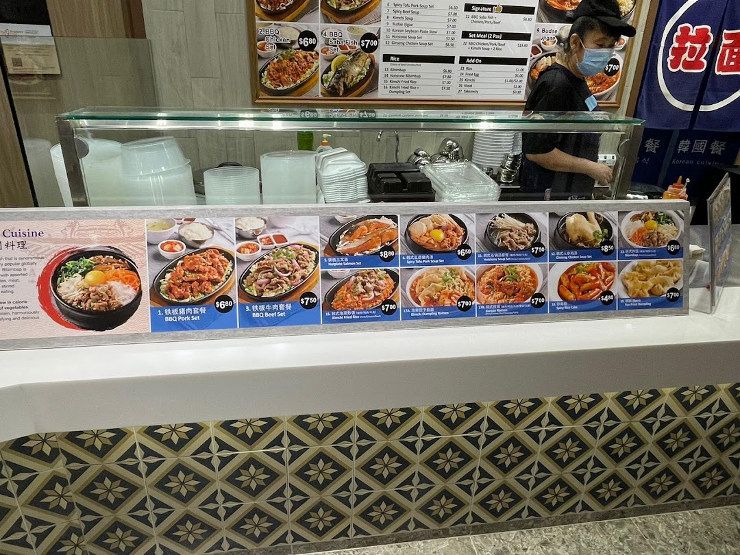 A vibrant counter at Kopitiam's Korean cuisine stall, showcasing a variety of delicious dishes and a staff member.