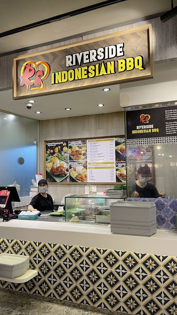 A bustling food stall counter at Riverside Indonesian BBQ, featuring staff and a detailed menu board.