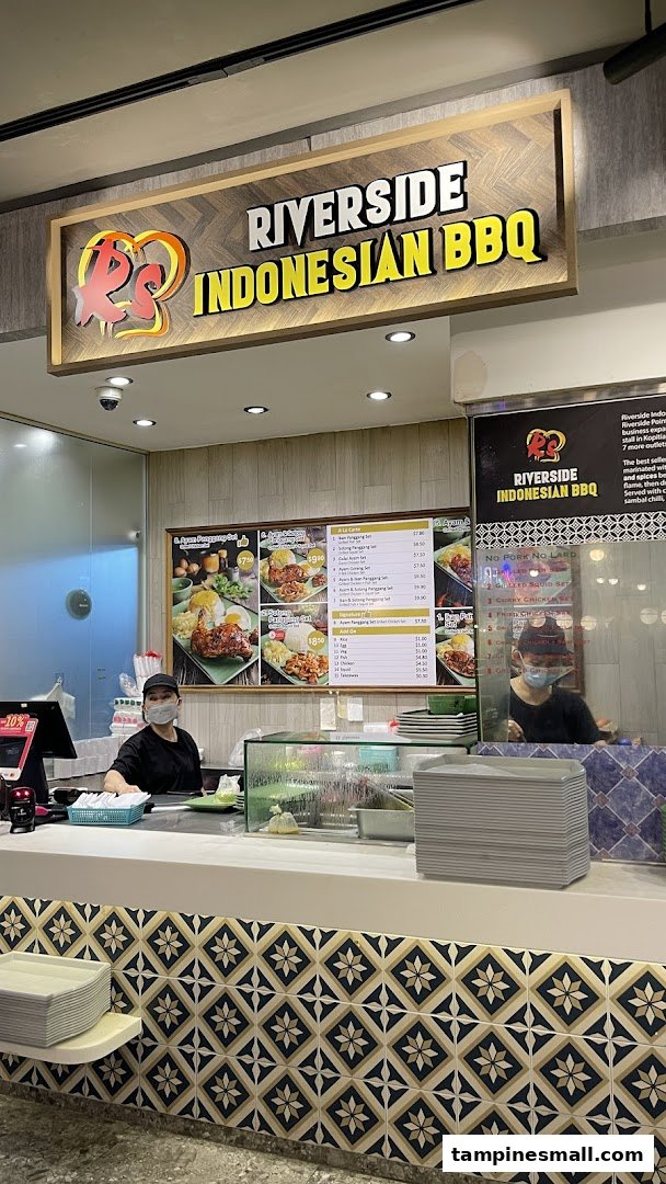 A bustling food stall counter at Riverside Indonesian BBQ, featuring staff and a detailed menu board.