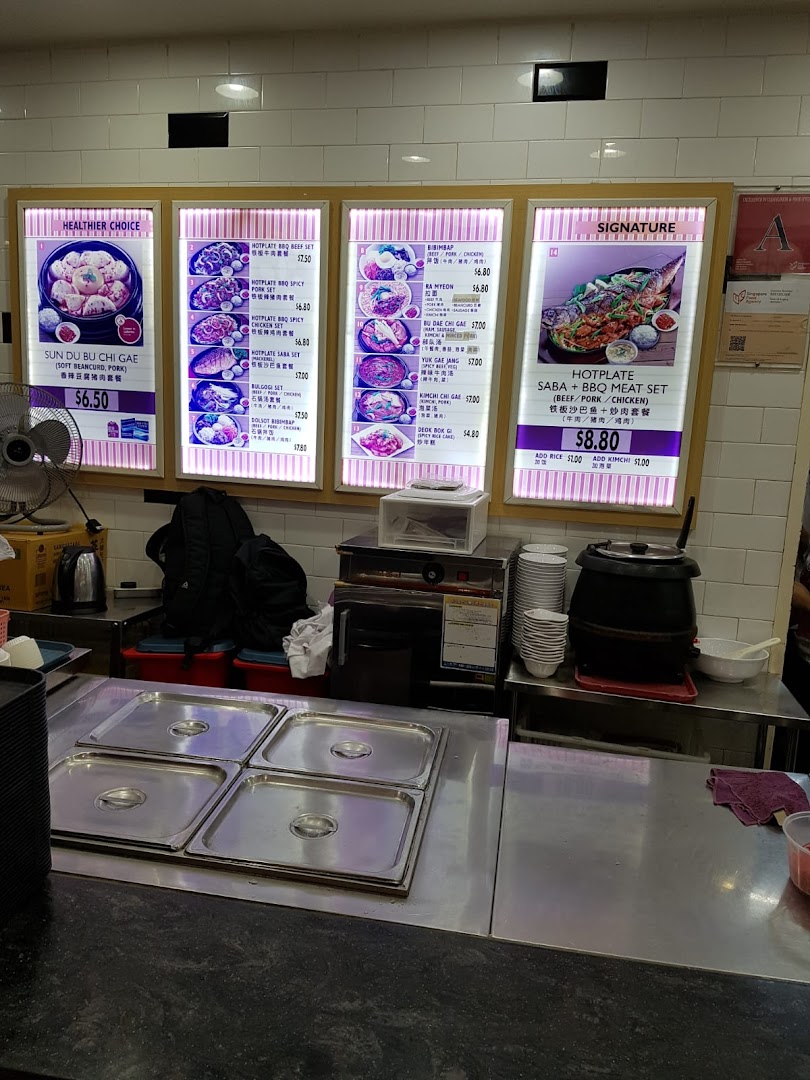 Brightly lit menu boards display a variety of Korean hotplate and stew dishes with prices at a food stall.