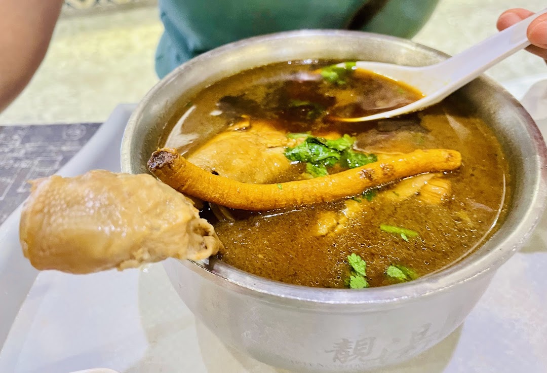 A close-up of a hearty bowl of herbal chicken soup with a chicken leg and a long root.