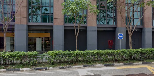 Exterior view of a building entrance with a yellow sign, trees, and a hedge along the roadside.