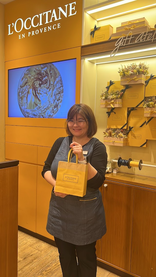 Smiling staff member holding a L'Occitane shopping bag inside the store.