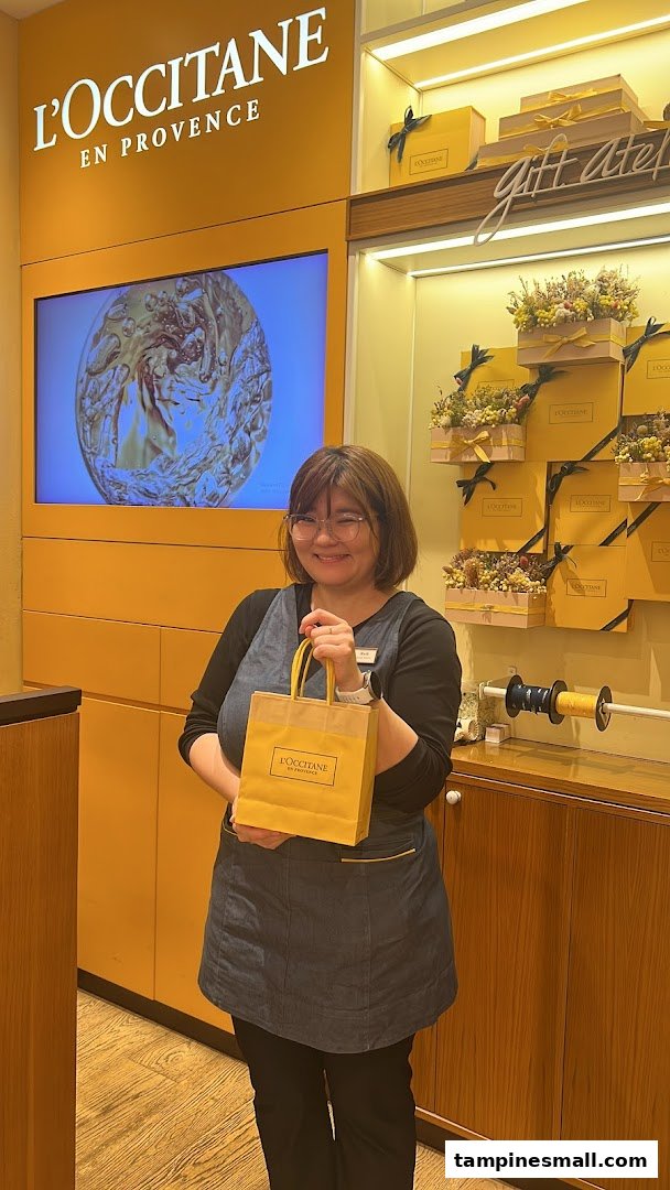 Smiling staff member holding a L'Occitane shopping bag inside the store.