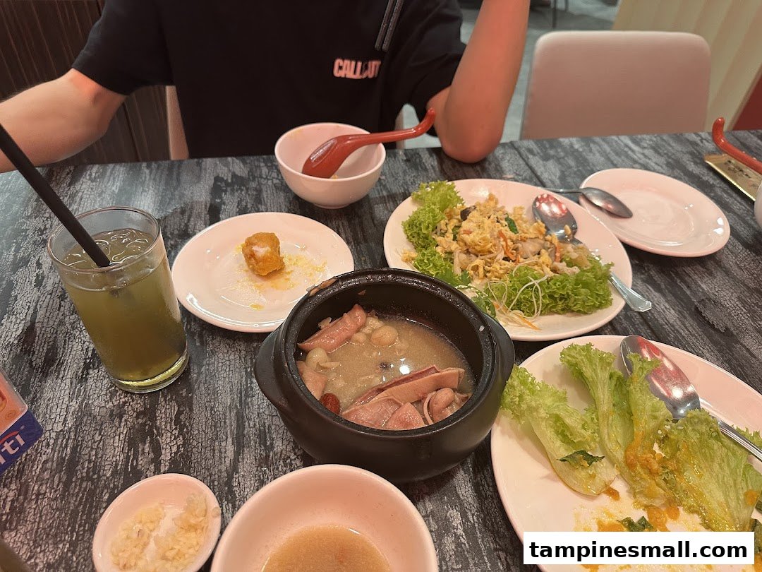 A table laden with various Chinese dishes, including soup, noodles, and a drink, with a person dining.