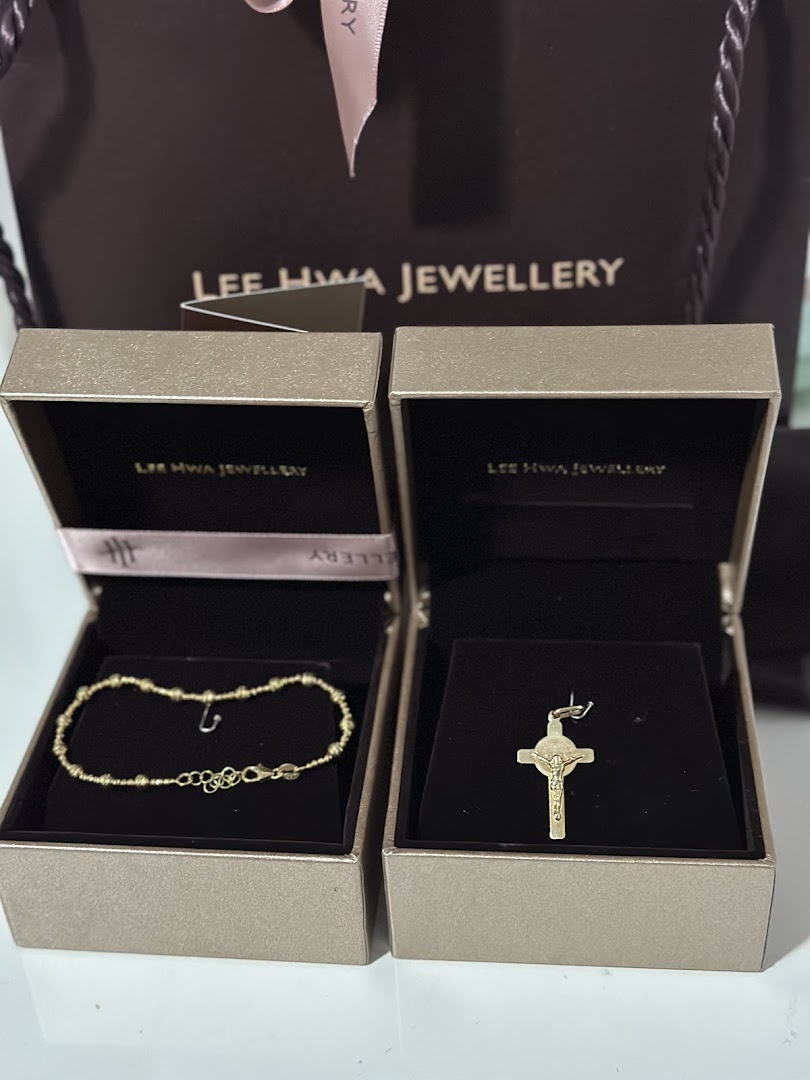 Two gold jewelry pieces, a bracelet and a cross pendant, displayed in Lee Hwa Jewellery boxes with a shopping bag.