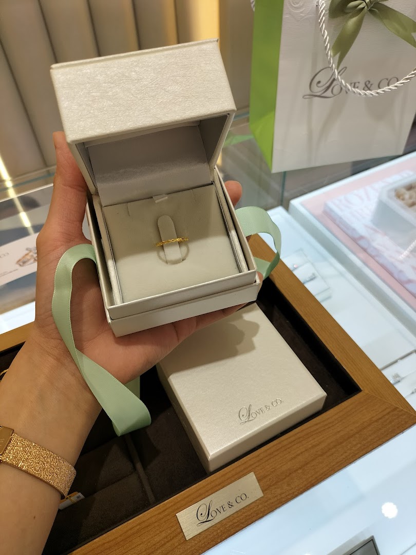 A hand holds an open Love & Co. ring box revealing a delicate gold ring, with branding visible.