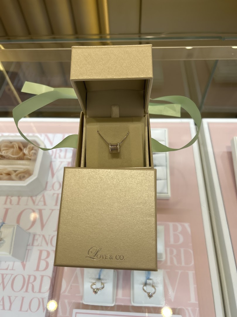 A delicate necklace displayed in an open golden box from Love & Co., alongside diamond rings.