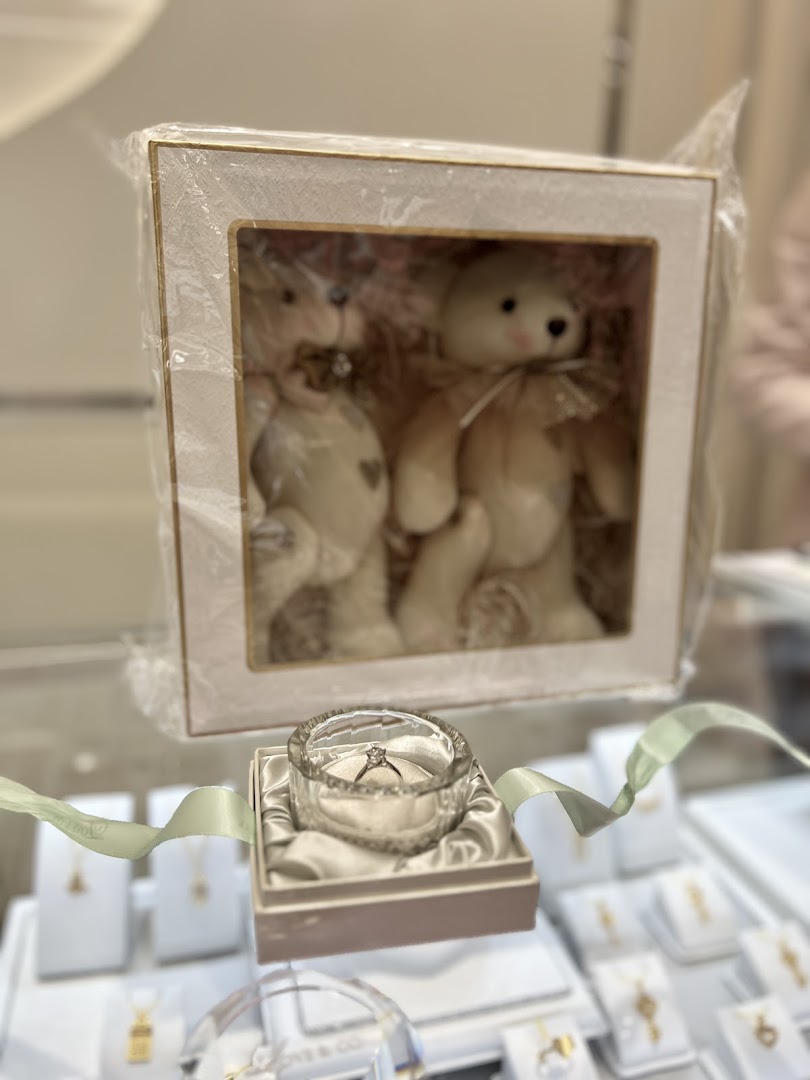 A close-up of a sparkling diamond engagement ring displayed with a box of charming teddy bears.