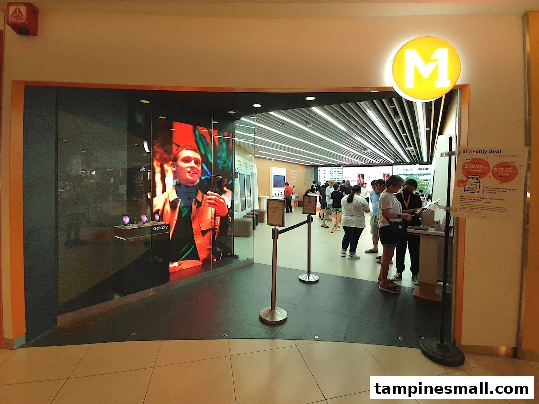The entrance of M1 Tampines Mall, a phone and internet service shop, with a large digital display and customers inside.