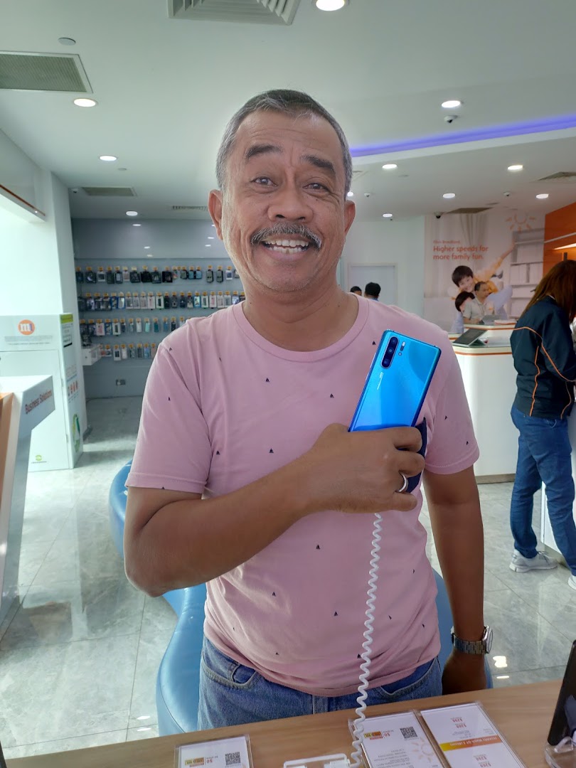 A smiling man holds a blue smartphone in a bright, modern phone shop.