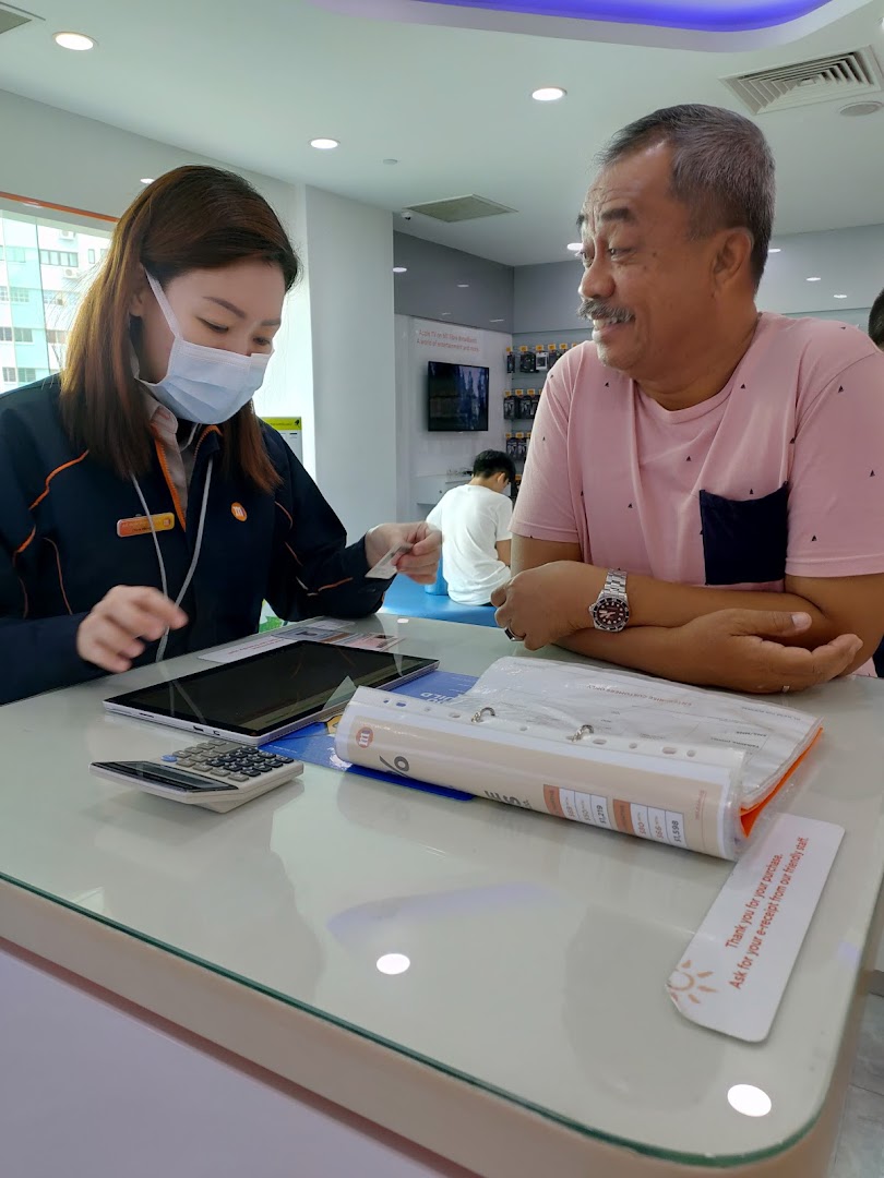A customer receives assistance from a masked staff member at an M1 phone and internet service counter.