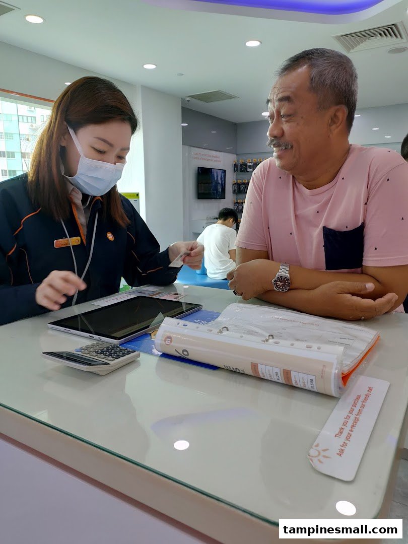 A customer receives assistance from a masked staff member at an M1 phone and internet service counter.