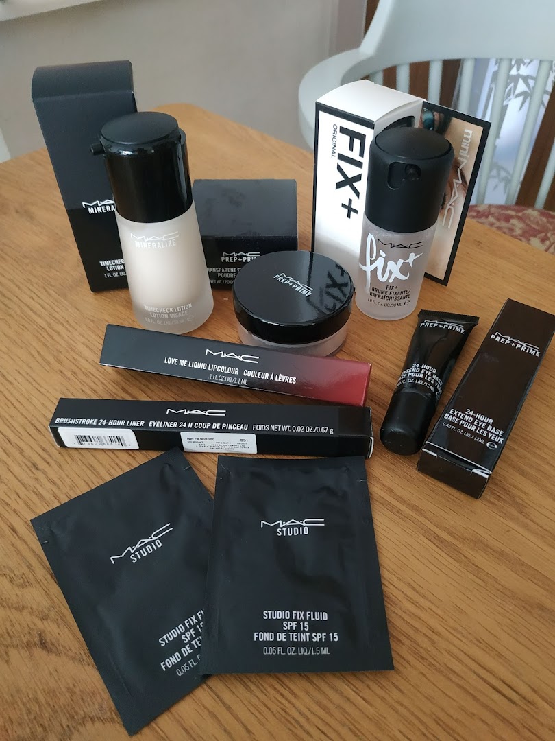 A collection of MAC Cosmetics makeup products, including foundations, primers, and samples, on a wooden table.