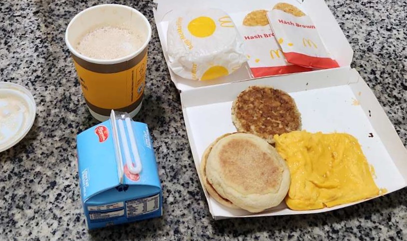 A McDonald's breakfast spread featuring coffee, milk, hash browns, and an open McMuffin meal.