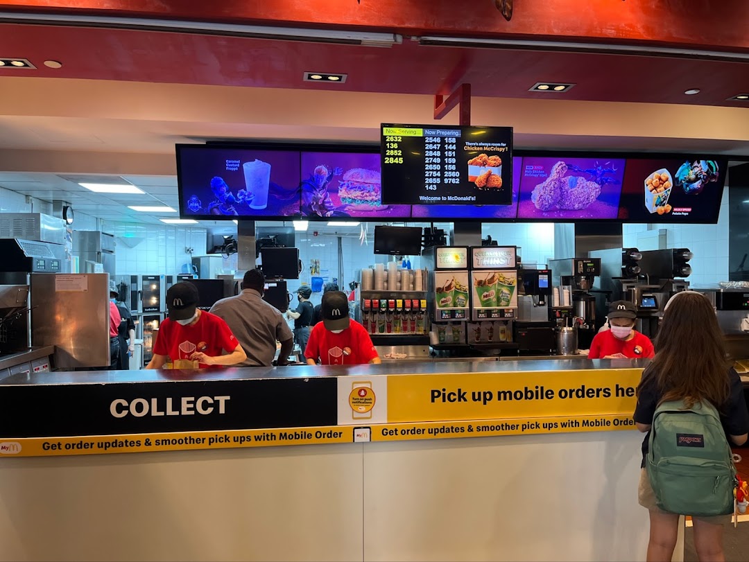 The image shows the bustling McDonald's order collection counter with staff and digital screens.