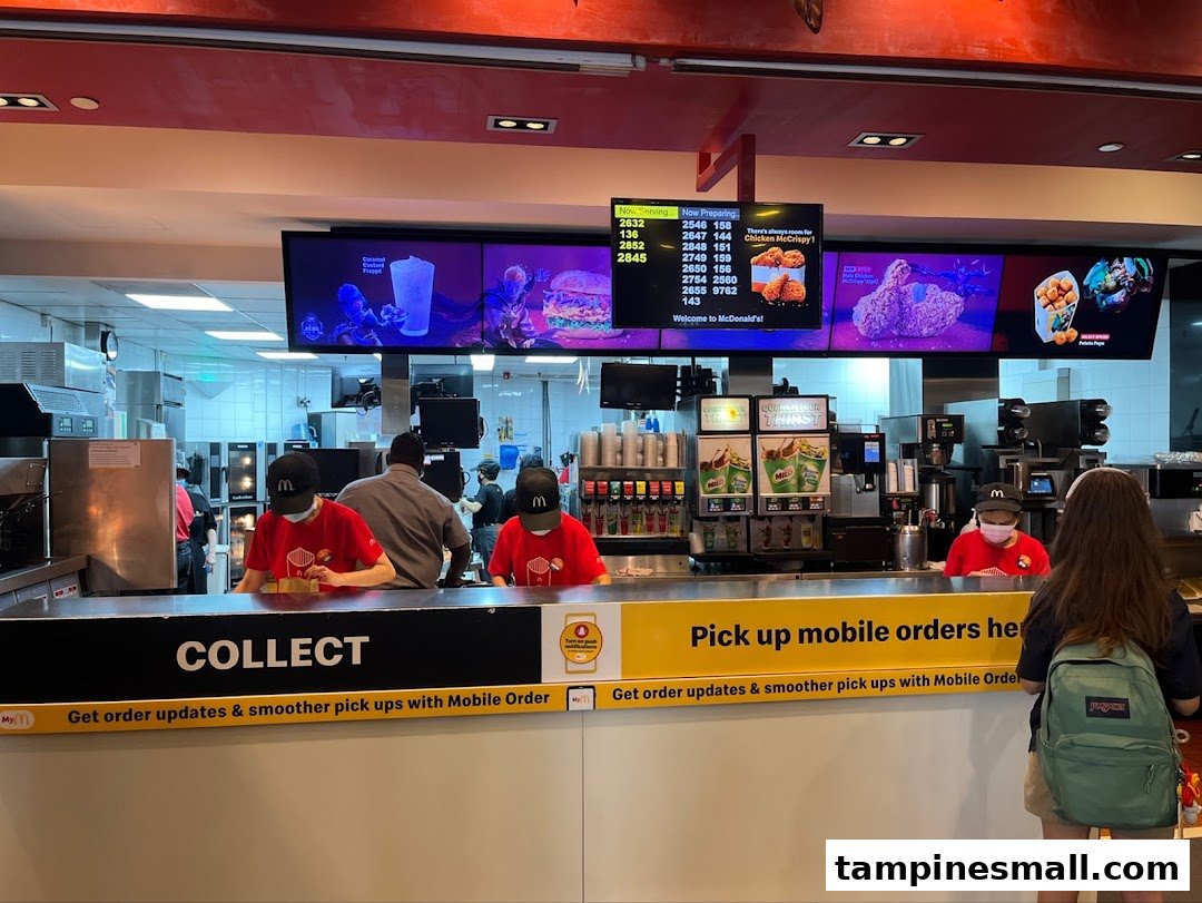 The image shows the bustling McDonald's order collection counter with staff and digital screens.
