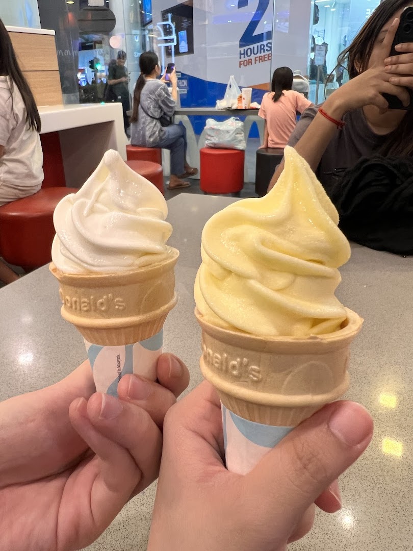 Two hands hold vanilla and pineapple soft-serve ice cream cones inside a McDonald's restaurant.