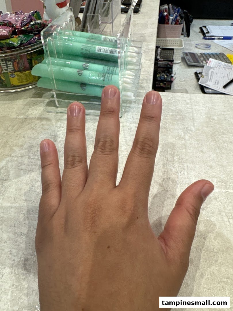 A hand with natural, well-maintained nails rests on a counter, with nail care products visible in the background.