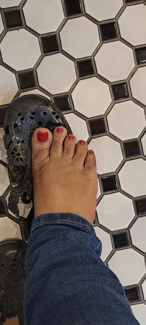 A close-up of a foot with vibrant red painted toenails and a decorative sandal on a black and white tiled floor.