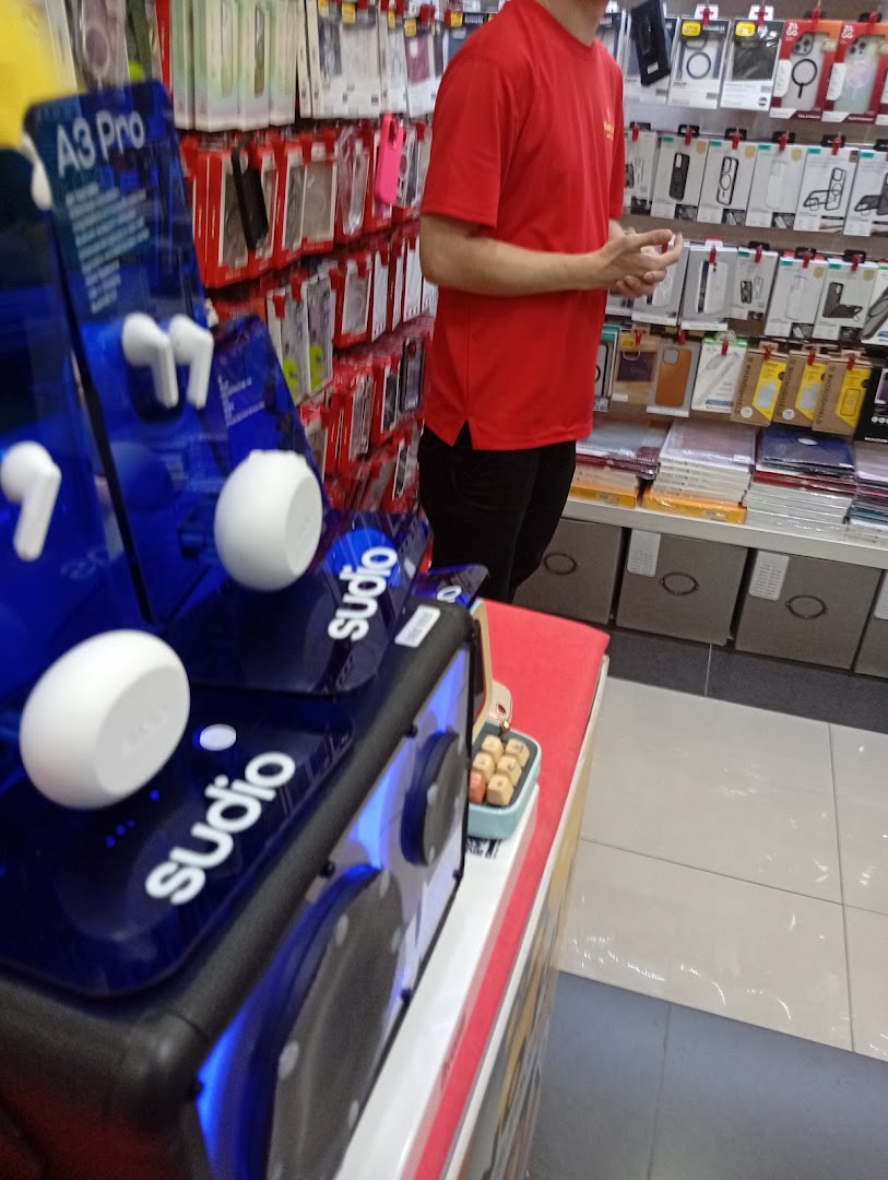 A person in a red shirt stands near a display of Sudio earbuds and various phone accessories in a tech shop.