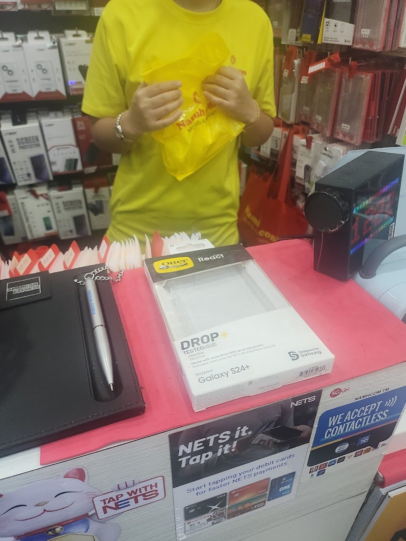 Interior of Namh.com, a mobile accessories shop, showing a staff member, product displays, and a payment counter.