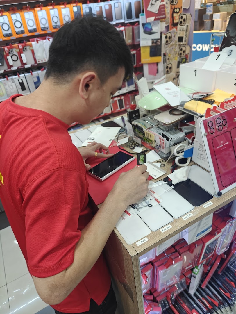 An employee in a red shirt applies a screen protector to a smartphone at a tech accessories shop.