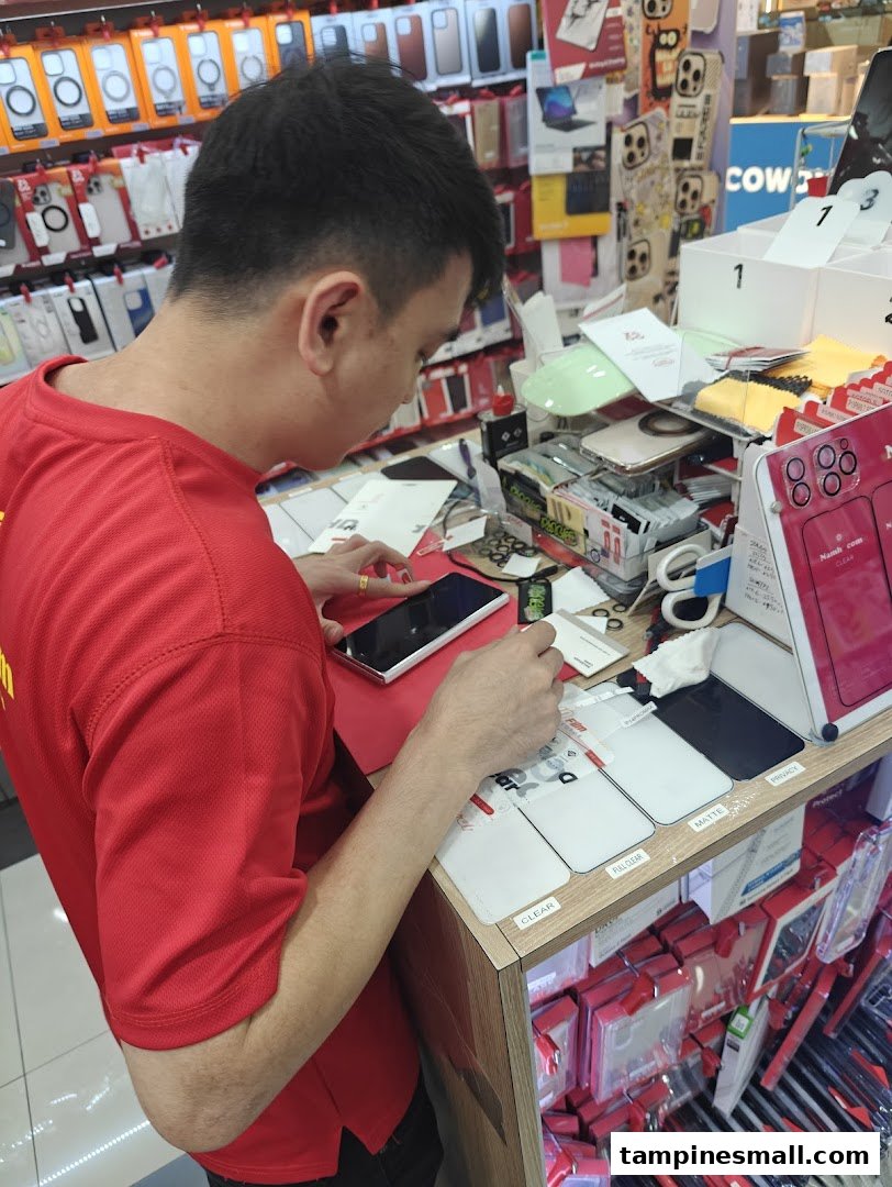 An employee in a red shirt applies a screen protector to a smartphone at a tech accessories shop.
