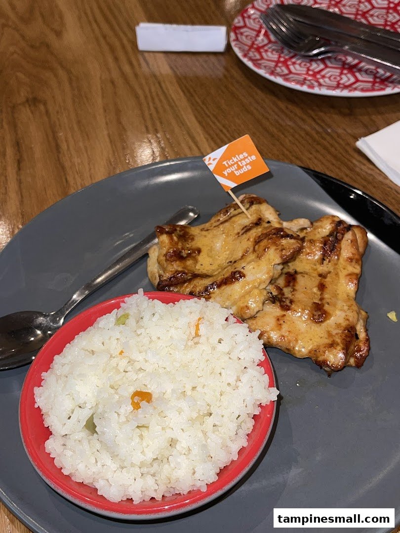 A plate of flame-grilled chicken with rice, featuring a 'Tickles your taste buds' flag, served on a grey plate.