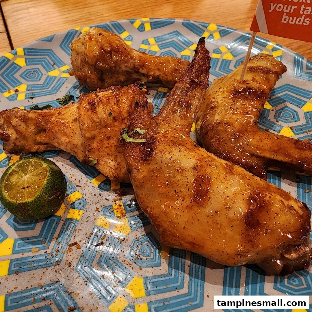Close-up of flame-grilled PERi-PERi chicken wings and drumsticks with a lime on a patterned plate.