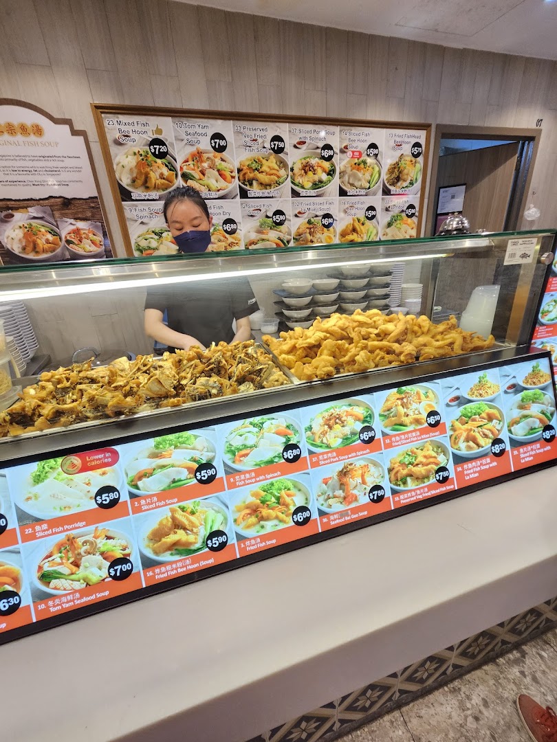 A food stall counter with a staff member, prepared fish dishes, and menu displays.