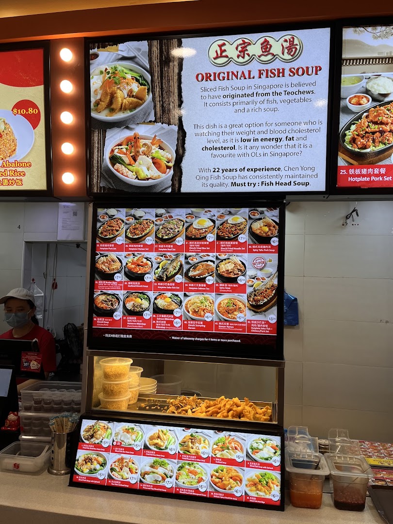 A food stall counter with digital menu boards displaying various fish soup dishes and a staff member.